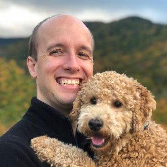 A bald white man in a black jacket holds an adorable doodle. Trees and other greenery are in the background.